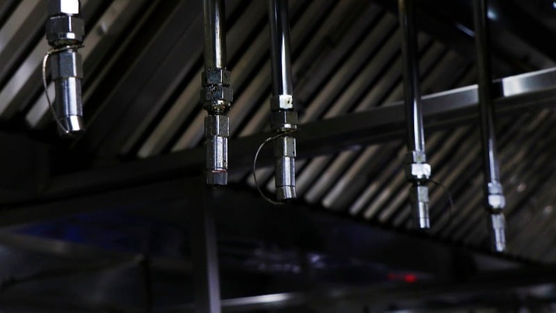 Featured image: Kitchen hood suppression system with pipes hanging down from the ceiling to put out fires - Read full post: How a Kitchen Hood Fire Suppression System Works